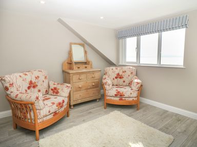 A room with two floral armchairs a wooden dresser with mirror a window with striped blinds and a white rug on a wooden floor at 11 Marine Terrace in Criccieth