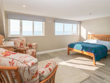 A bedroom with two floral armchairs a wooden bed with a blue blanket and windows overlooking the sea at 11 Marine Terrace in Criccieth