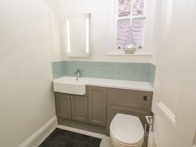 A bathroom with a white sink, grey cabinets, a toilet, a mirror, a window with frosted glass, and a decorative sailboat at 11 Marine Terrace in Criccieth