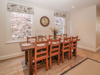 A dining room with a wooden table and eight chairs a clock on the wall and two windows at 11 Marine Terrace in Criccieth