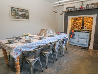 A dining room with a long table covered with a patterned tablecloth and set with bowls plates mugs and a vase of flowers and metal chairs next to a stone fireplace with a blue stove at 11 Marine Terrace in Criccieth