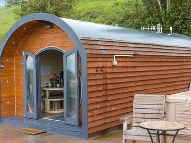 A wooden pod with open double doors showing a table with flowers inside and a wooden chair with a round table outside at Corndon Pod White Grit near Chirbury