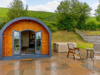 An outdoor patio with wooden chairs and a small table next to a curved wooden pod with open double doors at Corndon Pod White Grit near Chirbury