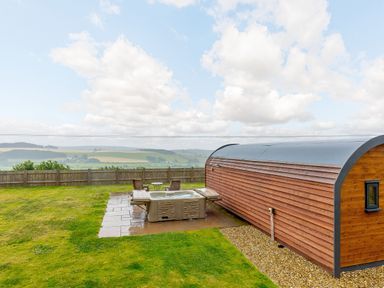 A wooden pod with a hot tub and chairs in a fenced grassy yard overlooking hills at Corndon Pod in White Grit near Chirbury