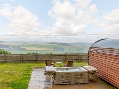 An outdoor patio with a hot tub wooden chairs a round metal table and a wooden fence with a field view at Corndon Pod White Grit near Chirbury