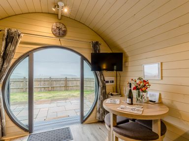 A wooden pod interior with a round glass door curtain a wall clock mounted tv a table with flowers and a bottle of wine at Corndon Pod White Grit near Chirbury