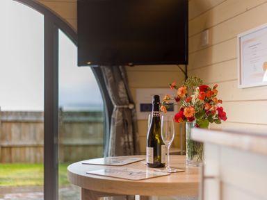 A table with a bottle, wine glass, and vase of flowers near a window and mounted TV at Corndon Pod in White Grit near Chirbury