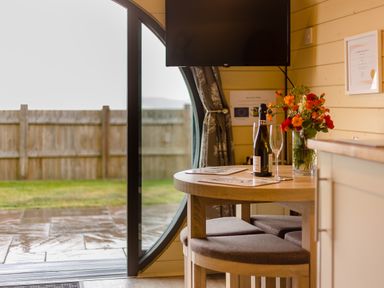 A wooden round table with a bottle, two glasses, and a vase with flowers near a round window and a wall-mounted TV at Corndon Pod in White Grit near Chirbury