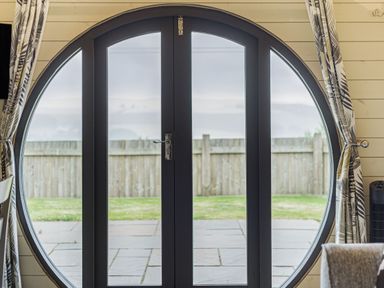 A circular black-framed glass door with patterned curtains and a view of a fenced yard at Stiperstones Pod in White Grit near Chirbury