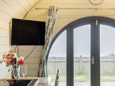 A room with a wall mounted TV flowers wine bottle and glasses on a table next to arched glass doors with curtains at Stiperstones Pod White Grit near Chirbury