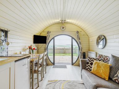 An interior room with a curved wooden ceiling a grey sofa with pillows a dining table with chairs a small kitchen area and a round sliding glass door at Stiperstones Pod in White Grit near Chirbury