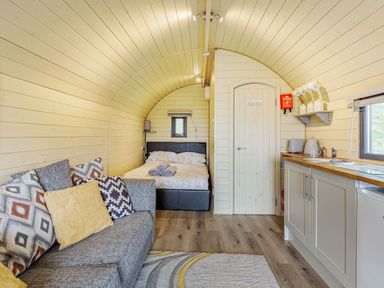 An interior with a sofa with cushions a bed wooden walls a door and a kitchenette at Stiperstones Pod in White Grit near Chirbury