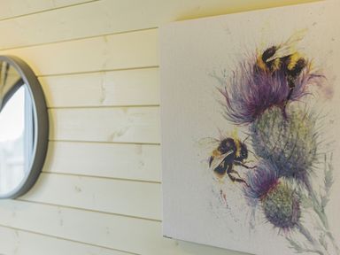 A wall with light-colored wood panels a round mirror and a painting of bees on thistle flowers at Stiperstones Pod in White Grit near Chirbury