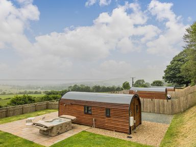 Wooden pods with curved roofs near a fenced yard with a hot tub and chairs in a rural setting at Stiperstones Pod in White Grit near Chirbury