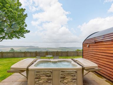 An outdoor hot tub on a patio next to a wooden structure with a table and two chairs on a lawn surrounded by a wooden fence at Stiperstones Pod in White Grit near Chirbury