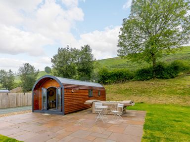 A small arched wooden pod with an open door on a stone patio next to a hot tub and two white chairs and a table at Stiperstones Pod in White Grit near Chirbury