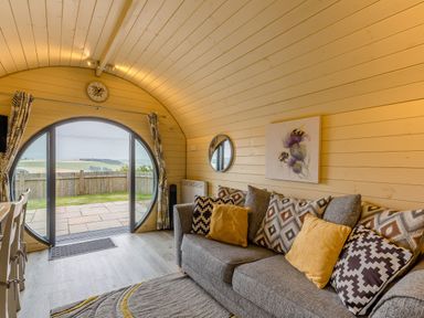 A living room with a gray sofa and patterned cushions near a circular glass door at Stiperstones Pod in White Grit near Chirbury