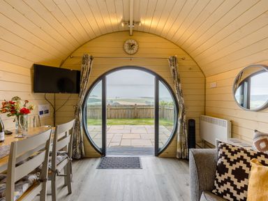 A dining area with a wooden table and chairs next to a mounted television and circular sliding door with curtains at Stiperstones Pod in White Grit near Chirbury