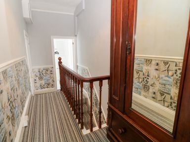 A hallway with striped carpet a wooden banister wallpaper with beach themed designs and a wooden wardrobe with mirror at Downlea in Looe