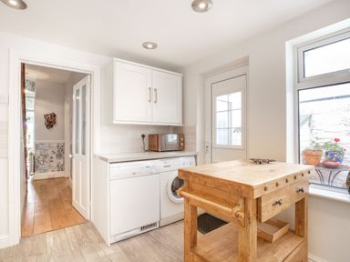 A kitchen with a wooden island, a microwave on the counter, dishwasher and washing machine under the counter, a door and window showing outdoor plants at Downlea in Looe