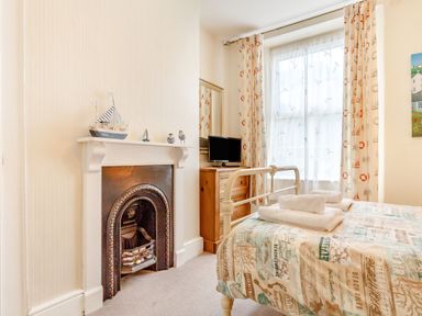 A bedroom with a metal bed, a wooden dresser, a small TV, patterned curtains, and a decorative fireplace at Downlea in Looe