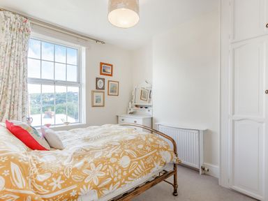 A bedroom with a bed covered with a yellow floral duvet a window with floral curtains a white dresser with a mirror and framed pictures on the wall at Downlea in Looe