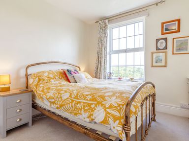 A bedroom with a metal bed frame and yellow patterned bedding a bedside table with a lamp and a window with floral curtains at Downlea in Looe