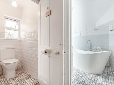 A bathroom with a white toilet and patterned floor tiles next to a room with a white freestanding bathtub and towels on a tiled floor at Downlea in Looe