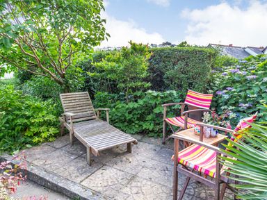 A garden patio with wooden lounge chair two striped chairs and a small table with plants and cups surrounded by bushes at Downlea in Looe