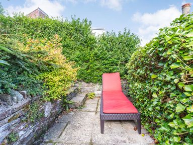 A red cushioned lounge chair on a stone patio surrounded by green hedges and plants at Downlea in Looe
