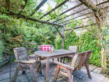 An outdoor seating area with a wooden table and four chairs under a pergola with climbing plants at Downlea in Looe