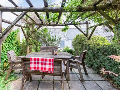 An outdoor patio with a wooden table and chairs under a wooden pergola with plants at Downlea in Looe