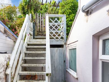 A white wooden outdoor staircase with a railing next to a house wall with windows and plants at Downlea in Looe