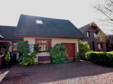A small house with a sloped roof bushes a bench and a gravel driveway at Swallow Lodge in Amble