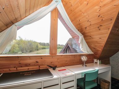 A wooden attic room with a white desk, green chair, small mirror, toiletries, and a large window with curtains at Swallow Lodge in Amble