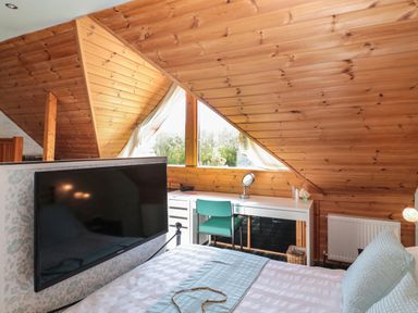 A bedroom with a bed a mounted TV and a desk with a chair under a slanted wooden ceiling at Swallow Lodge in Amble