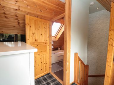 A wooden door open to a bathroom with a bathtub under a skylight next to a wooden staircase with patterned wallpaper at Swallow Lodge in Amble