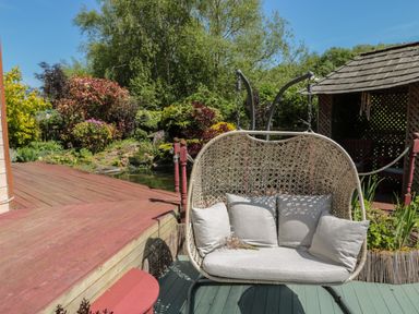 An outdoor garden area with a hanging wicker loveseat with cushions near a wooden deck and a small pond at Swallow Lodge in Amble