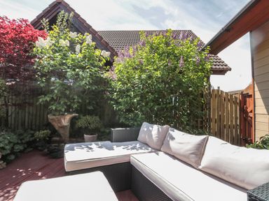 An outdoor patio with a wicker sofa with white cushions and surrounding plants at Swallow Lodge in Amble