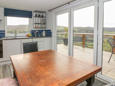 A kitchen with a wooden table and black chair near glass doors leading to a deck overlooking hills at Haka Lodge in Devil's Bridge