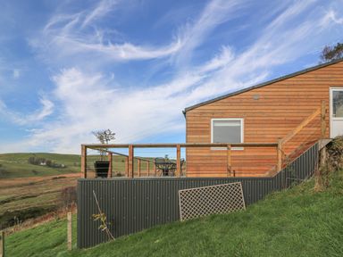 An outdoor deck with table and chairs attached to a wooden building with railing on a grassy hillside at Haka Lodge in Devil's Bridge