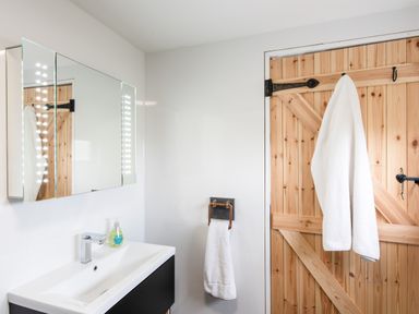 A bathroom with a white sink and mirror lighted with bulbs wooden door with two white towels hanging on hooks at Haka Lodge in Devil's Bridge