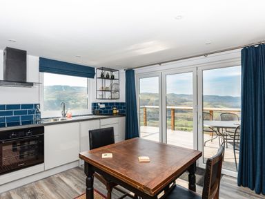 A kitchen with white cabinets and blue tile backsplash a wooden table with black chairs a window and glass doors opening to a balcony with chairs and a table at Haka Lodge in Devil's Bridge