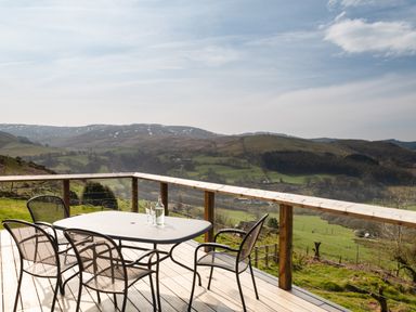 An outdoor deck with a table and four chairs overlooking a green landscape with hills at Haka Lodge in Devil's Bridge