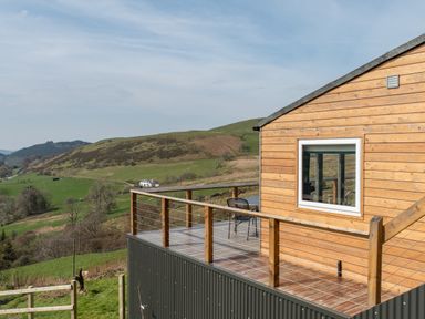 A wooden cabin with a small balcony and two chairs overlooking green hills at Haka Lodge in Devil's Bridge