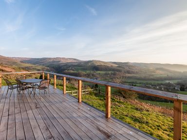 An outdoor wooden deck with a table and four chairs overlooking grassy hills and valleys at Haka Lodge in Devil's Bridge