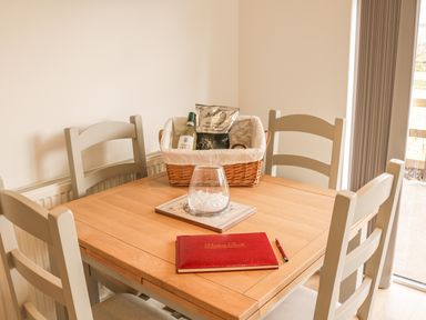 A wooden dining table with four chairs a wicker basket with snacks and a wine bottle a glass bowl and a red visitors book with a pen at Upham View in Woodbury