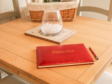 A wooden table with a red visitors book and pen a glass container with decorative crystals and a wicker basket at Upham View in Woodbury