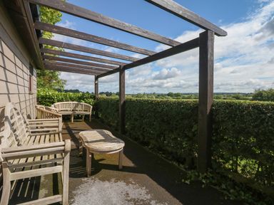 An outdoor patio with wooden benches and a table under a wooden pergola with hedges and a countryside view at Upham View in Woodbury