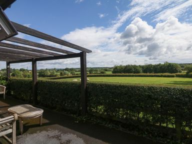 A patio with wooden furniture and a pergola overlooking a hedge and green fields at Upham View in Woodbury